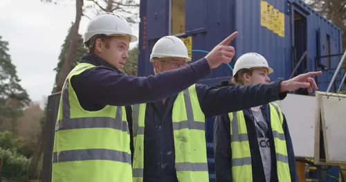 Three men in high vis jackets and helmets pointing at a house